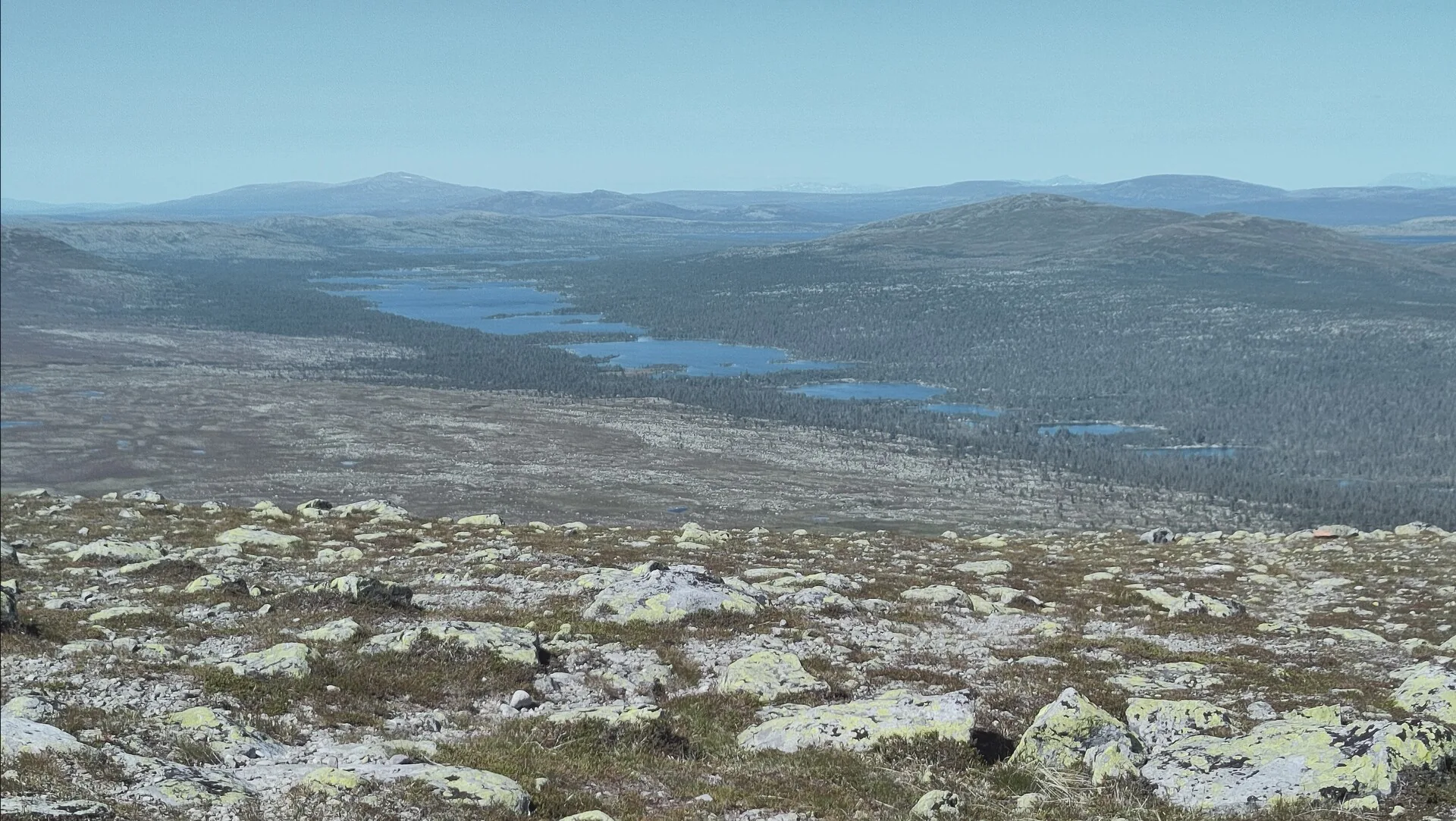 Blick von einer kargen Hochfläche mit Flechtensteinen über eine Seenkette und Wald bis zu fernen Bergrücken in Schweden